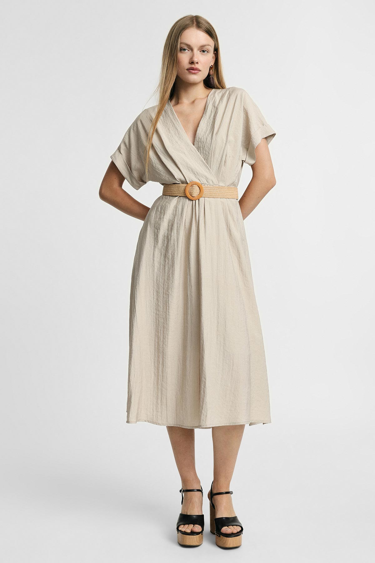 Model in beige midi wrap dress with short sleeves and woven belt, black platform sandals, standing against a white background