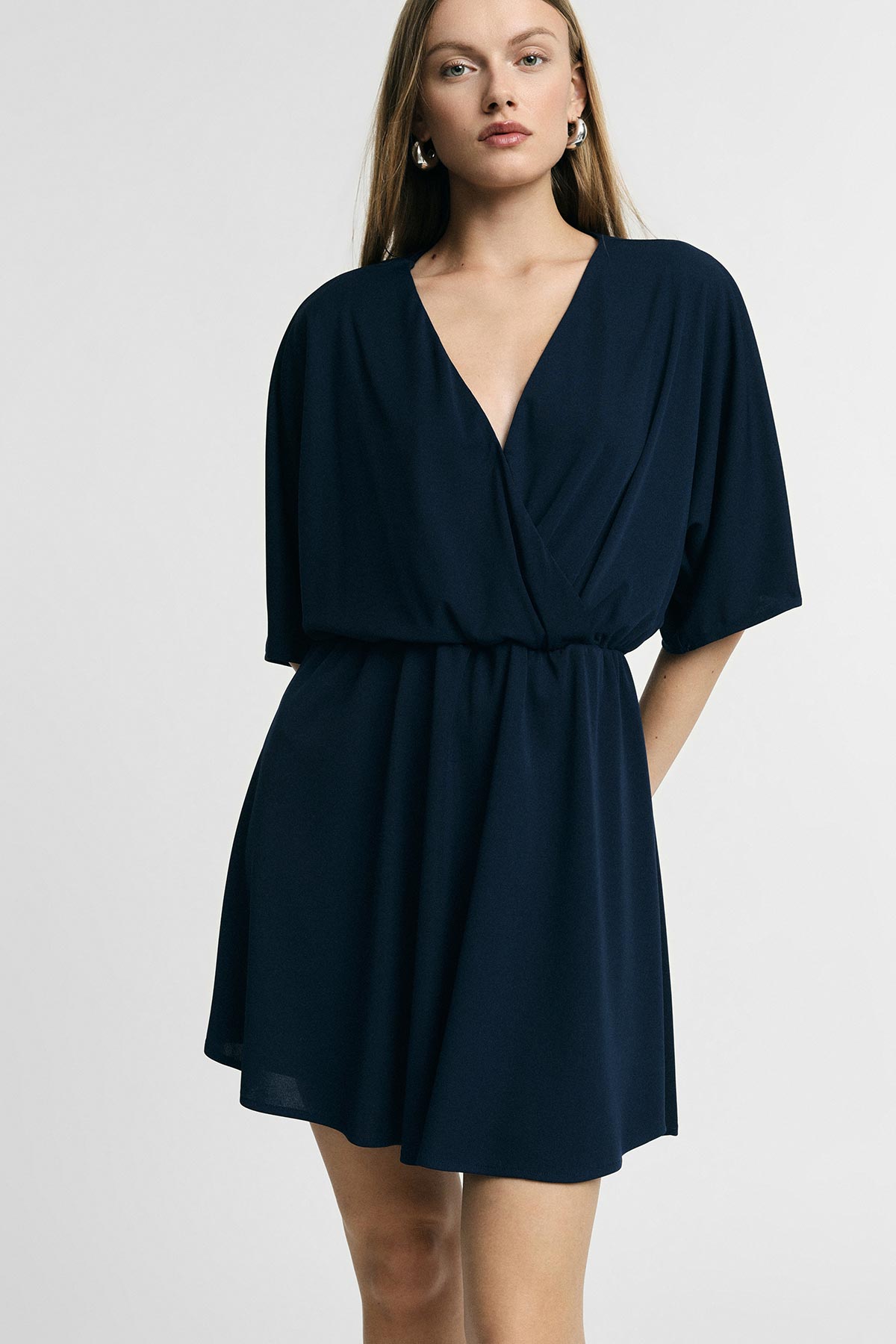 Model wearing navy V‑neck wrap dress with short kimono sleeves and elasticated waist, standing against light backdrop