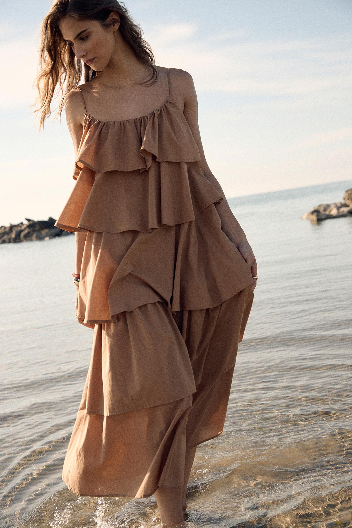 Young woman in a tiered brown summer dress walking barefoot in shallow sea with rocky coastline in soft light