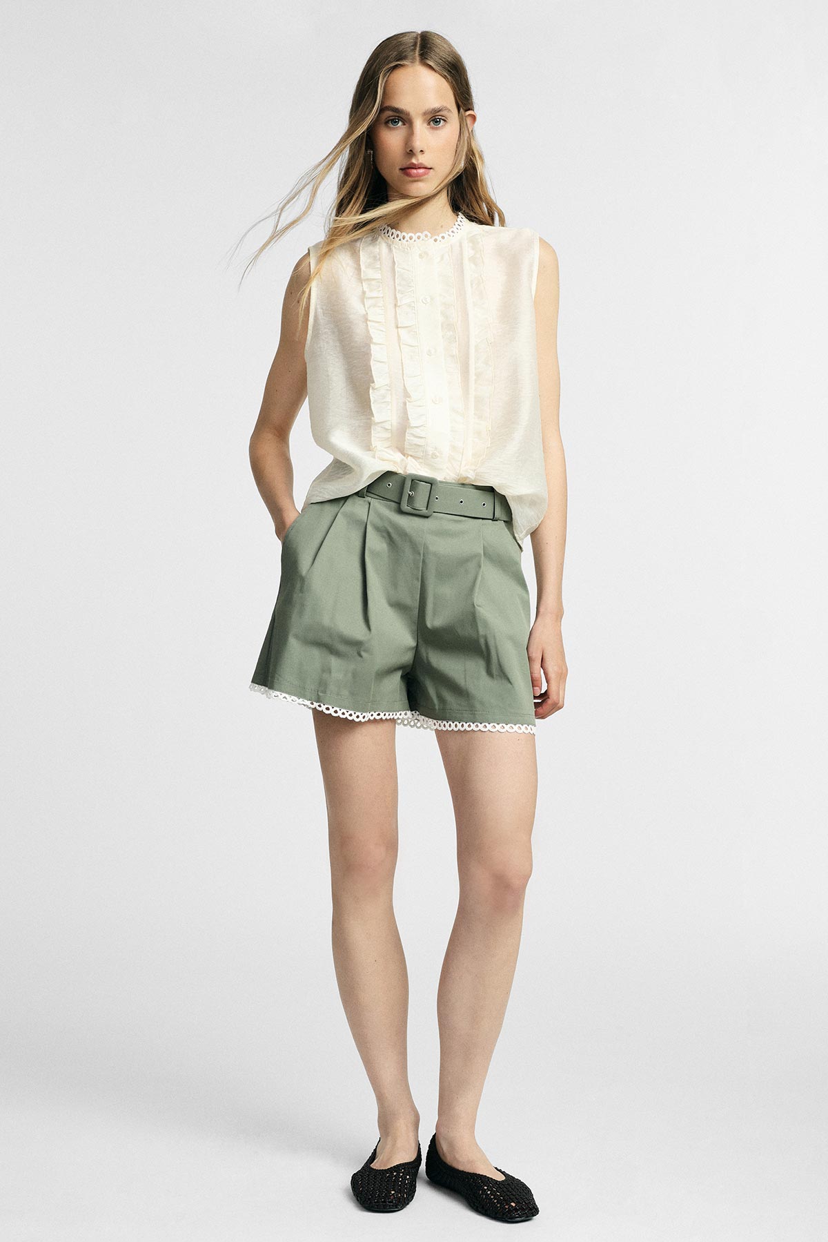 Female model wearing cream sleeveless ruffled blouse and high-waist sage green belted shorts with lace trim and black flats