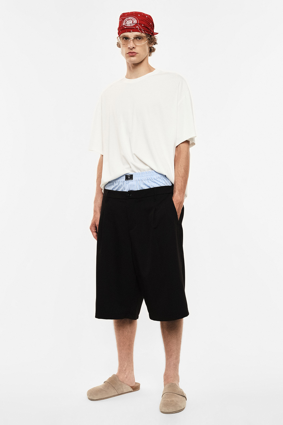 Man wearing oversized white T-shirt, black knee-length shorts, red bandana, clear glasses and beige slippers