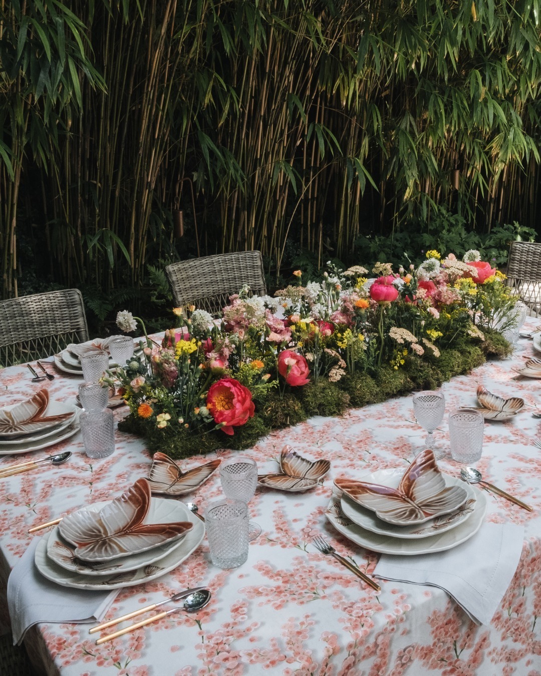 Outdoor dining table with wicker chairs, pink-peony and wildflower moss centrepiece, patterned pink tablecloth and elegant place settings