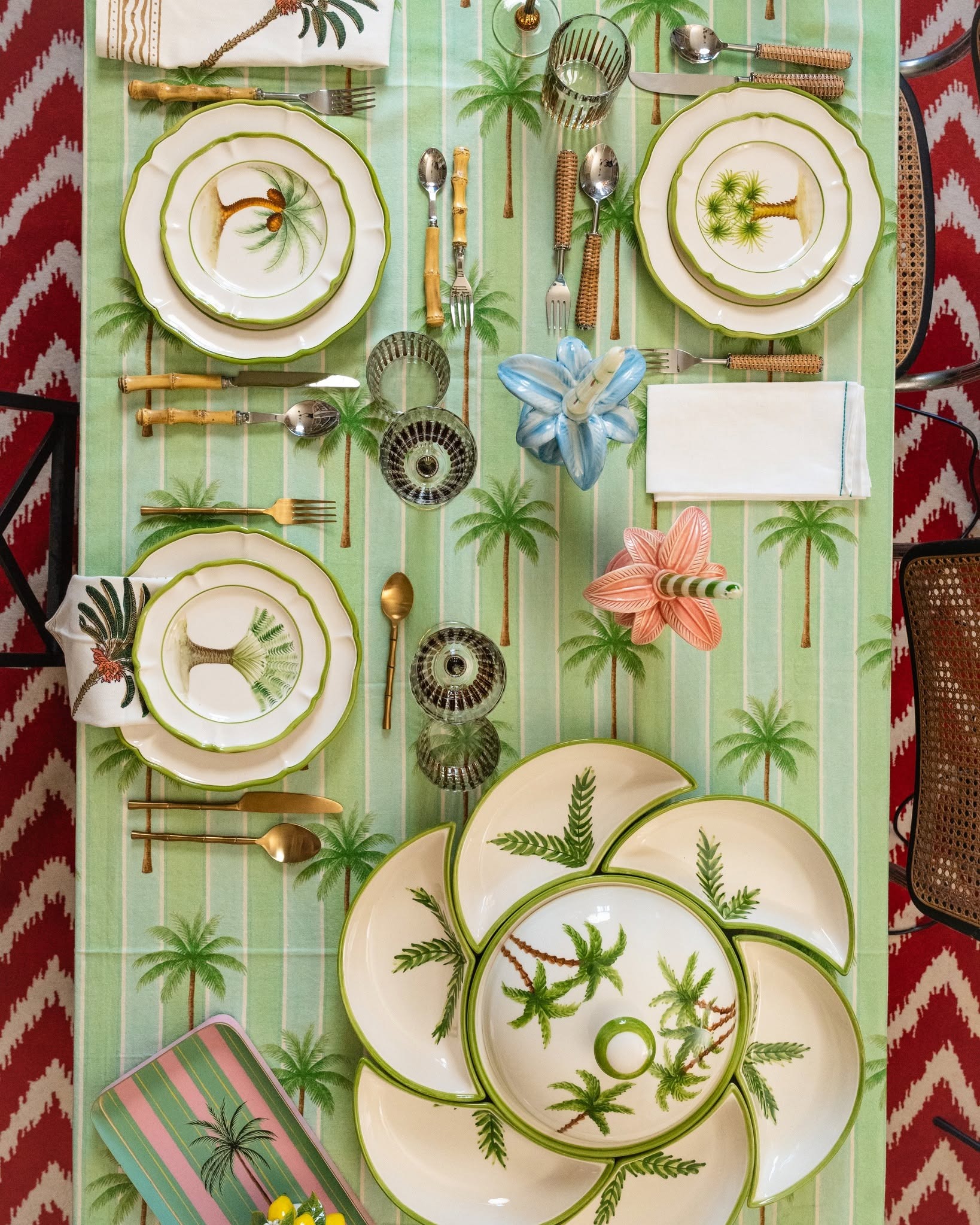 Top-down view of a tropical-themed dining table with palm-print plates, green striped tablecloth, glassware, cutlery and decorative napkins
