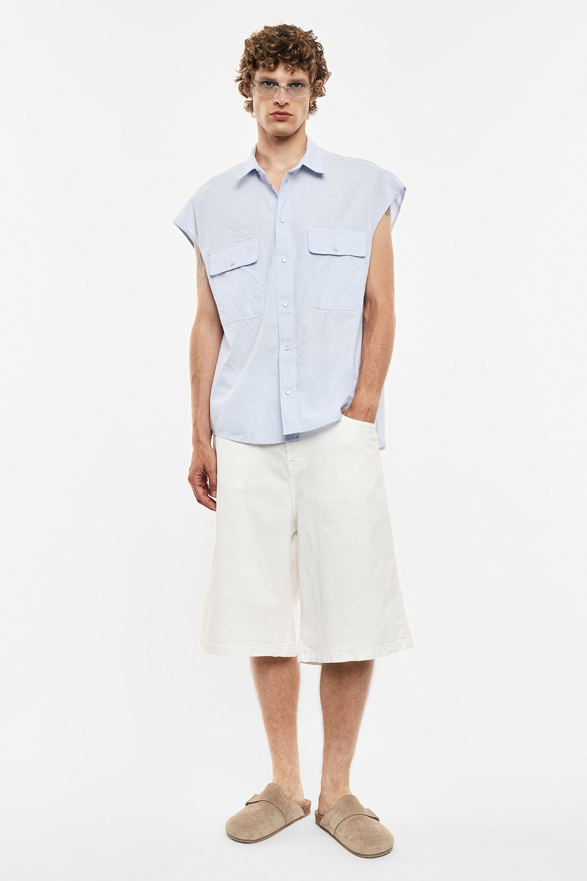 Young man in light blue sleeveless shirt, white wide-leg knee-length shorts and beige slip-on shoes, wearing glasses