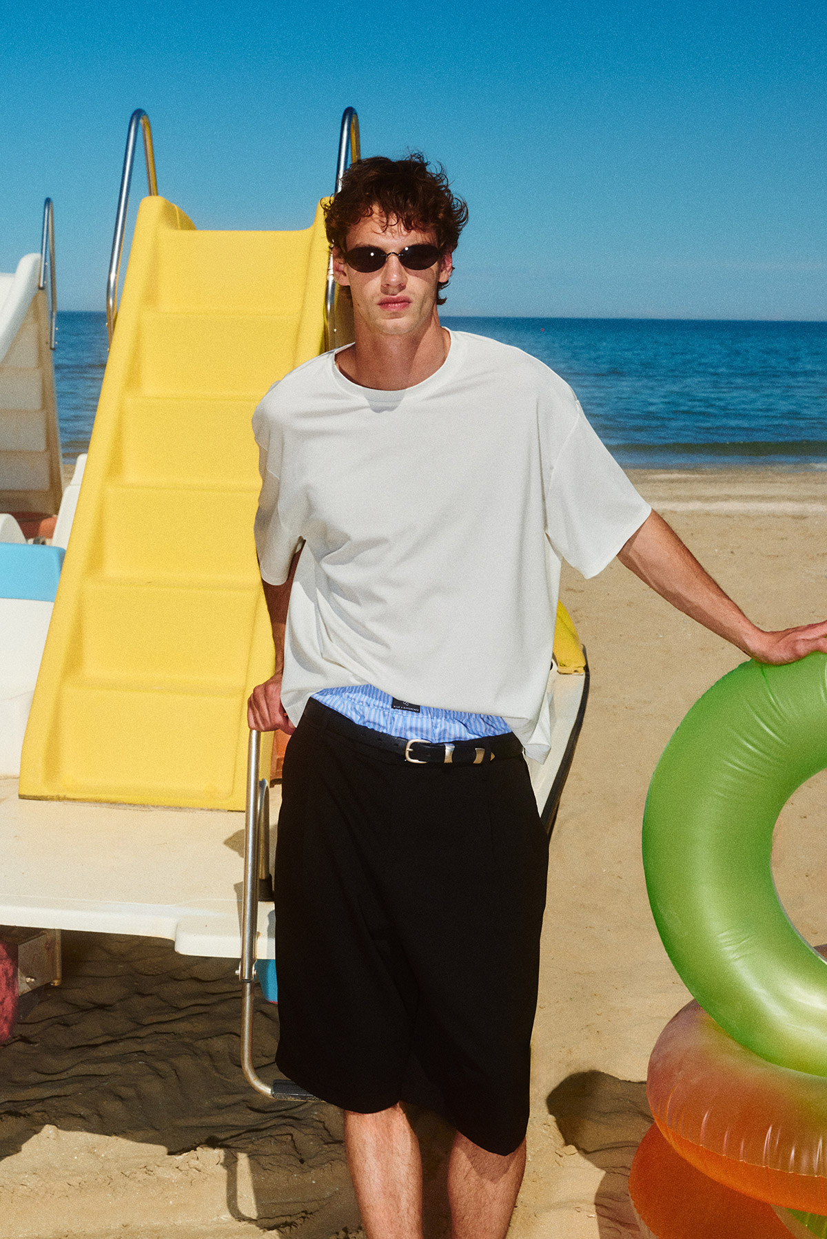 Young man in white T-shirt and sunglasses standing on a sandy beach by a yellow slide and green inflatable ring, sea in the background