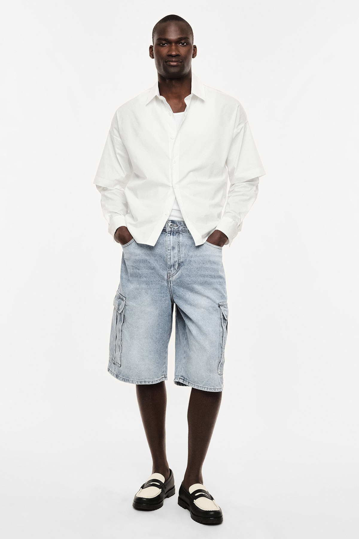 Man wearing white button-up shirt, light denim cargo shorts and black-and-white loafers, hands in pockets, standing