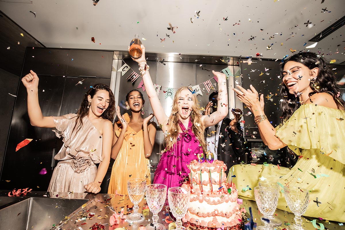 Group of diverse women cheering at a colourful birthday party with confetti, multi-tier cake and champagne