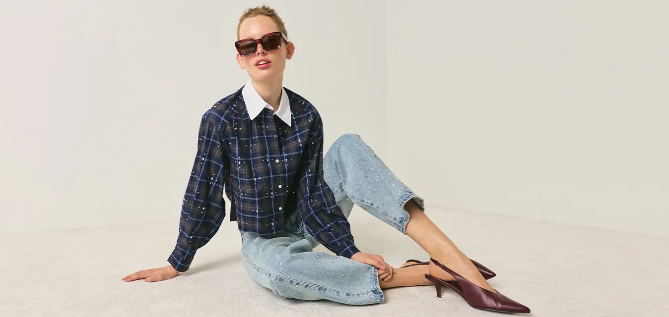 Donna con occhiali da sole, camicia scura a quadri, jeans azzurri chiari e tacchi viola seduta sul pavimento contro uno sfondo neutro semplice.
