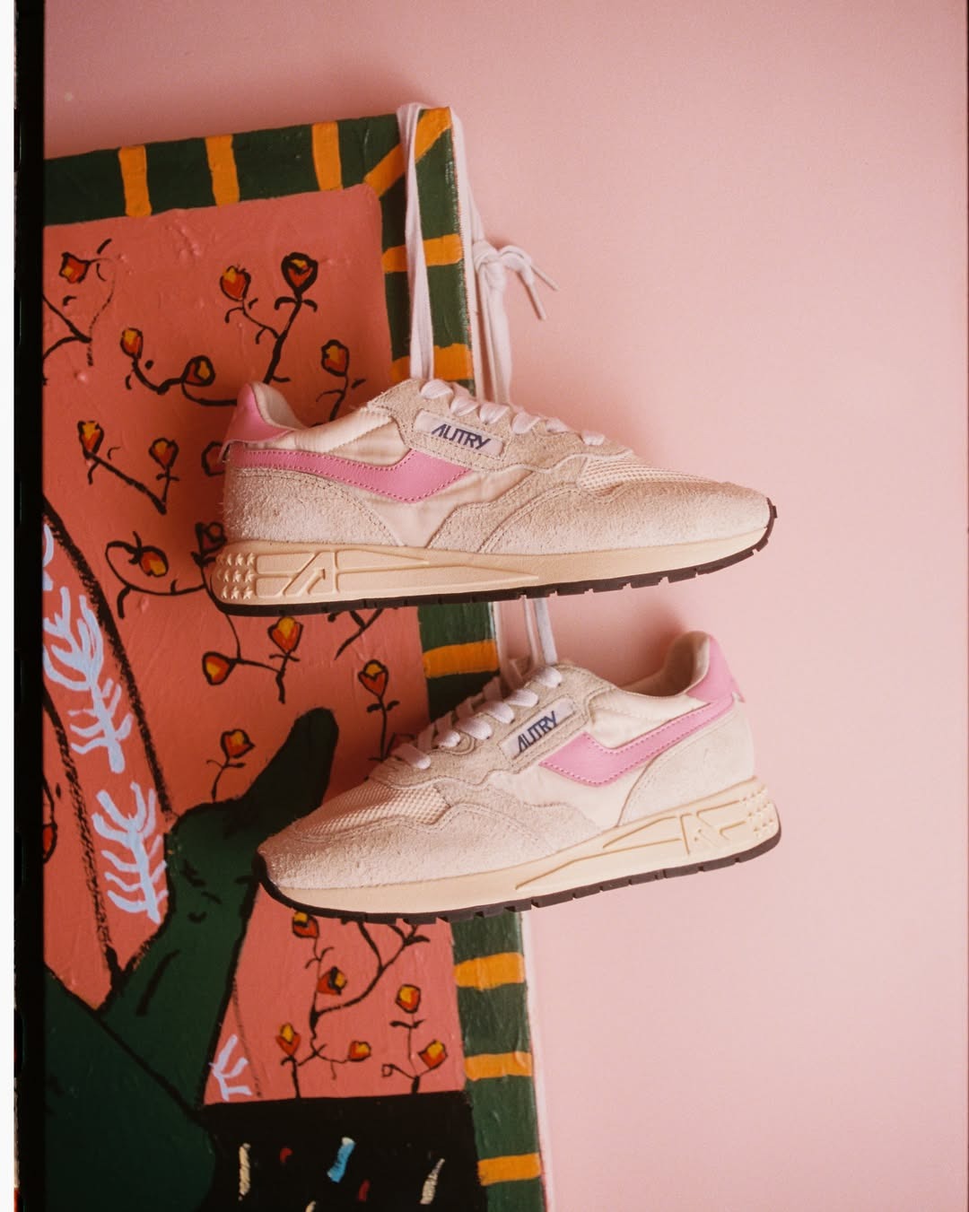 Beige and pink Autry suede trainers hanging by laces against a pink wall and floral-painted wooden panel