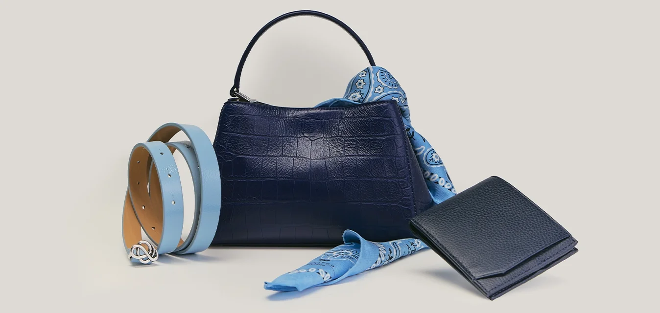 Borsa blu navy con sciarpa a fantasia blu, cintura doppia azzurra chiara e sabbia, e portafoglio blu navy disposti su superficie beige.