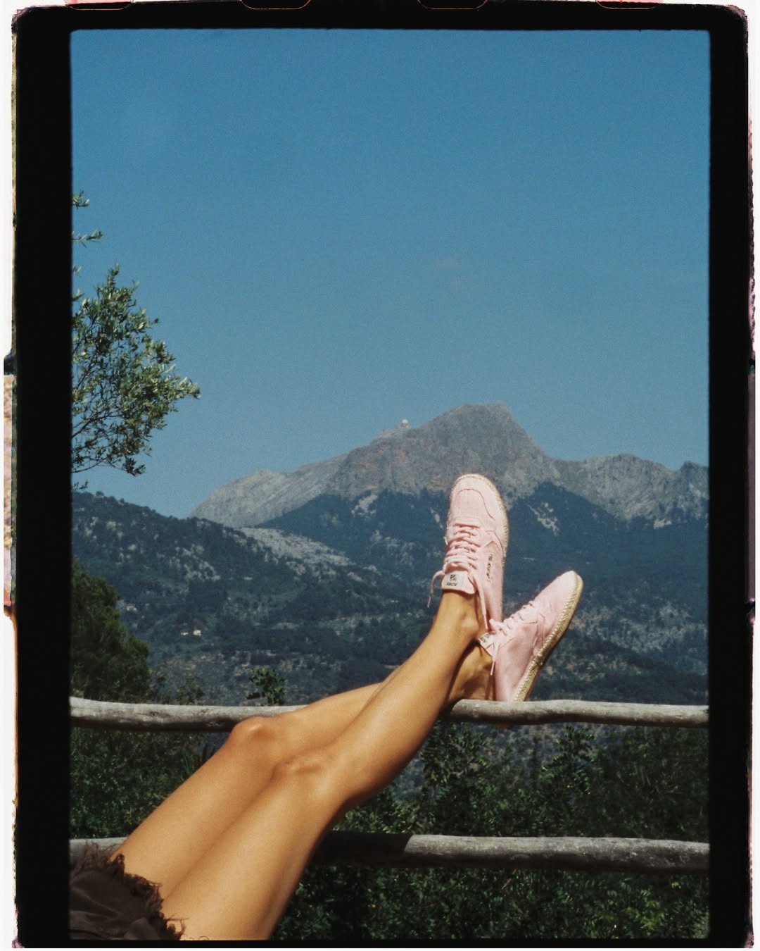 Tanned legs in pink trainers resting on a wooden fence with mountains and blue sky in the background