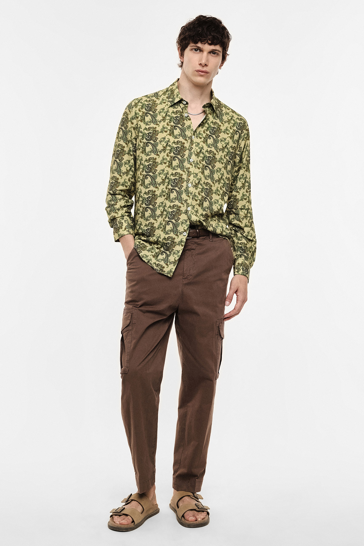 Male model wearing green paisley long-sleeve shirt, brown cargo trousers and tan sandals in a casual studio portrait