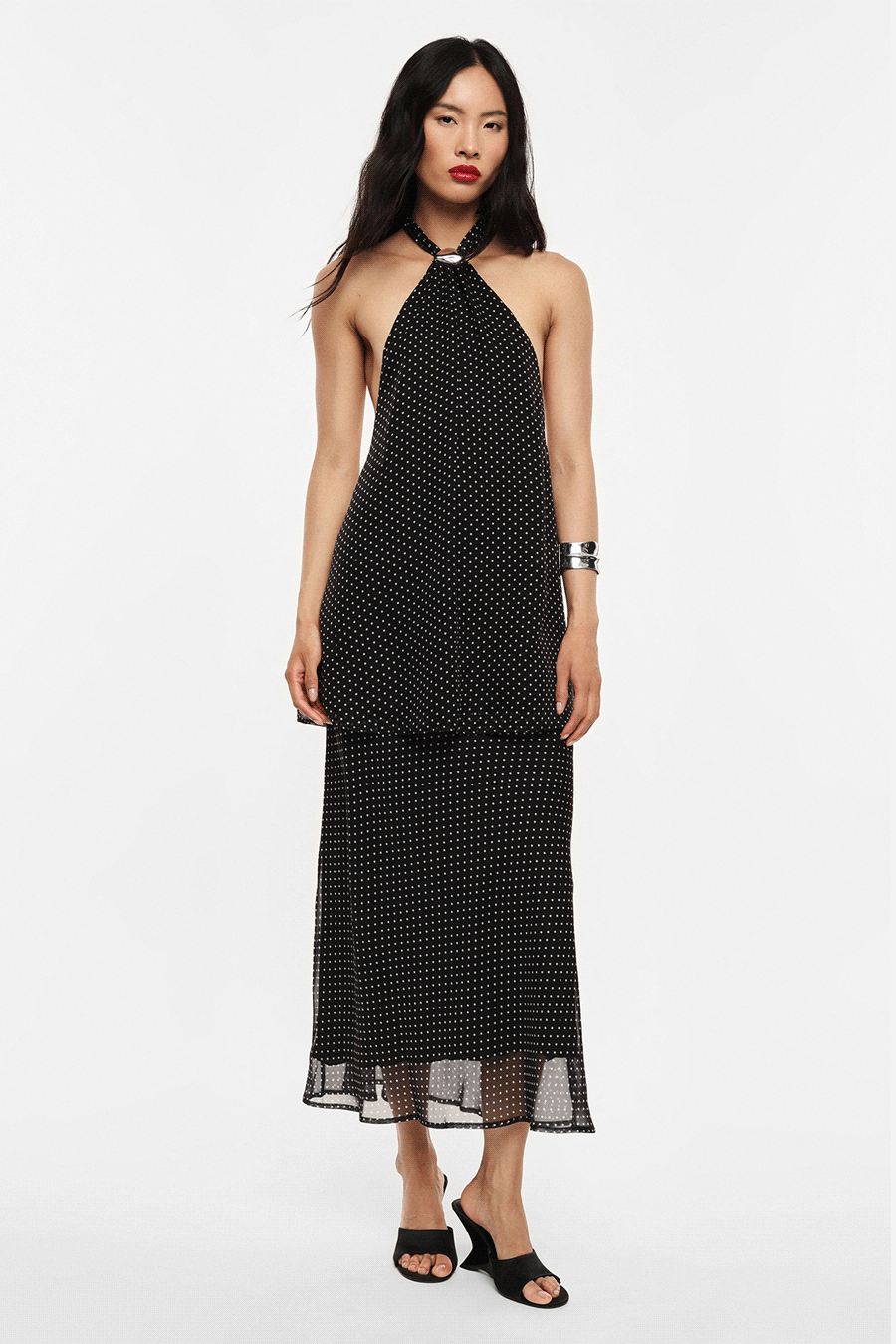 Woman modelling black polka-dot halter backless midi dress with tie-neck, sheer hem, black sandals, silver cuff bracelet; front and back views