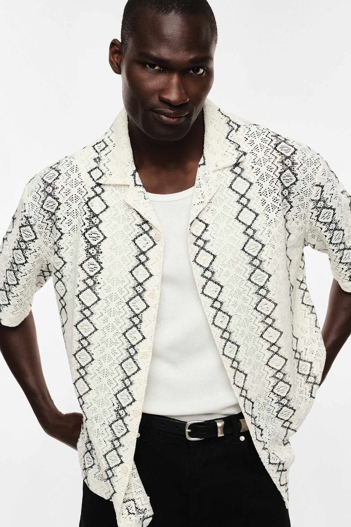 Male model wearing cream short-sleeve patterned knit shirt over a white vest, black jeans with belt, posing against a white background