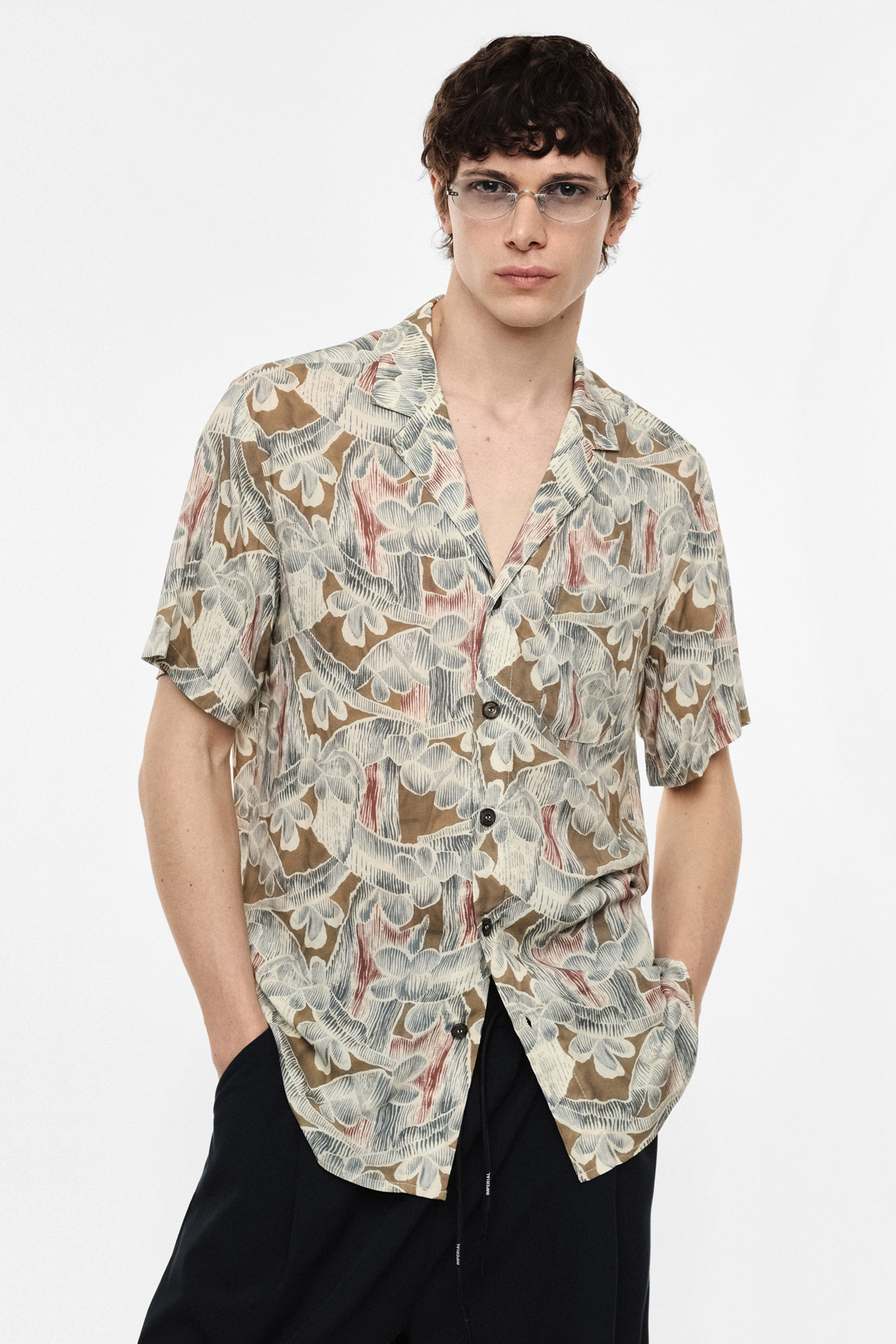 Young man wearing beige patterned short-sleeve summer shirt and clear glasses, hands in pockets against plain white background