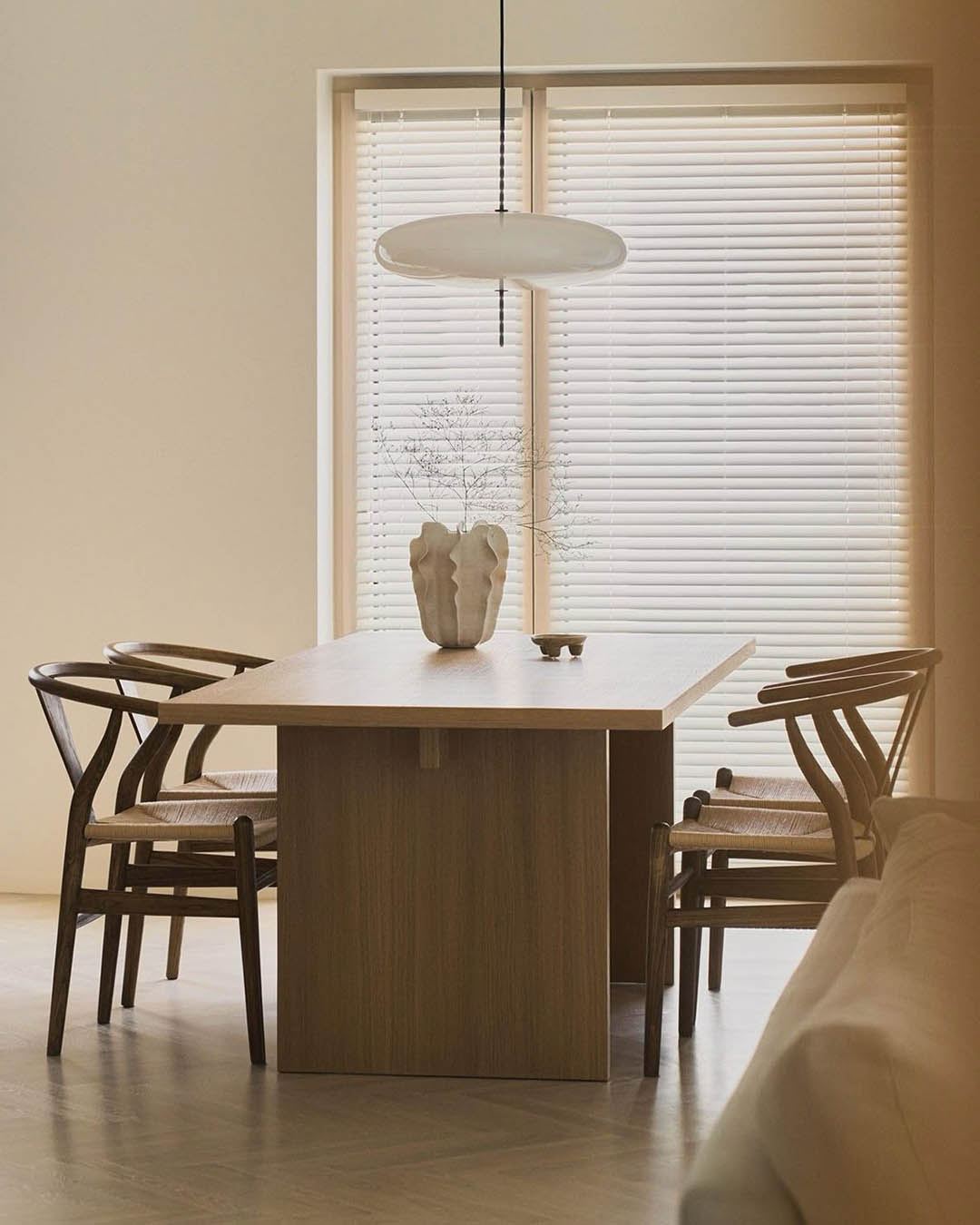 Minimalist dining area with wooden table, four woven-seat chairs, sculptural vase on table and round pendant light by closed window blinds