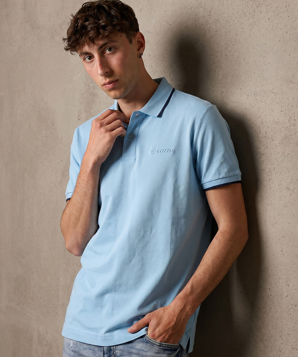 Young man wearing light blue polo shirt with navy trim, standing against a textured concrete wall in a relaxed pose
