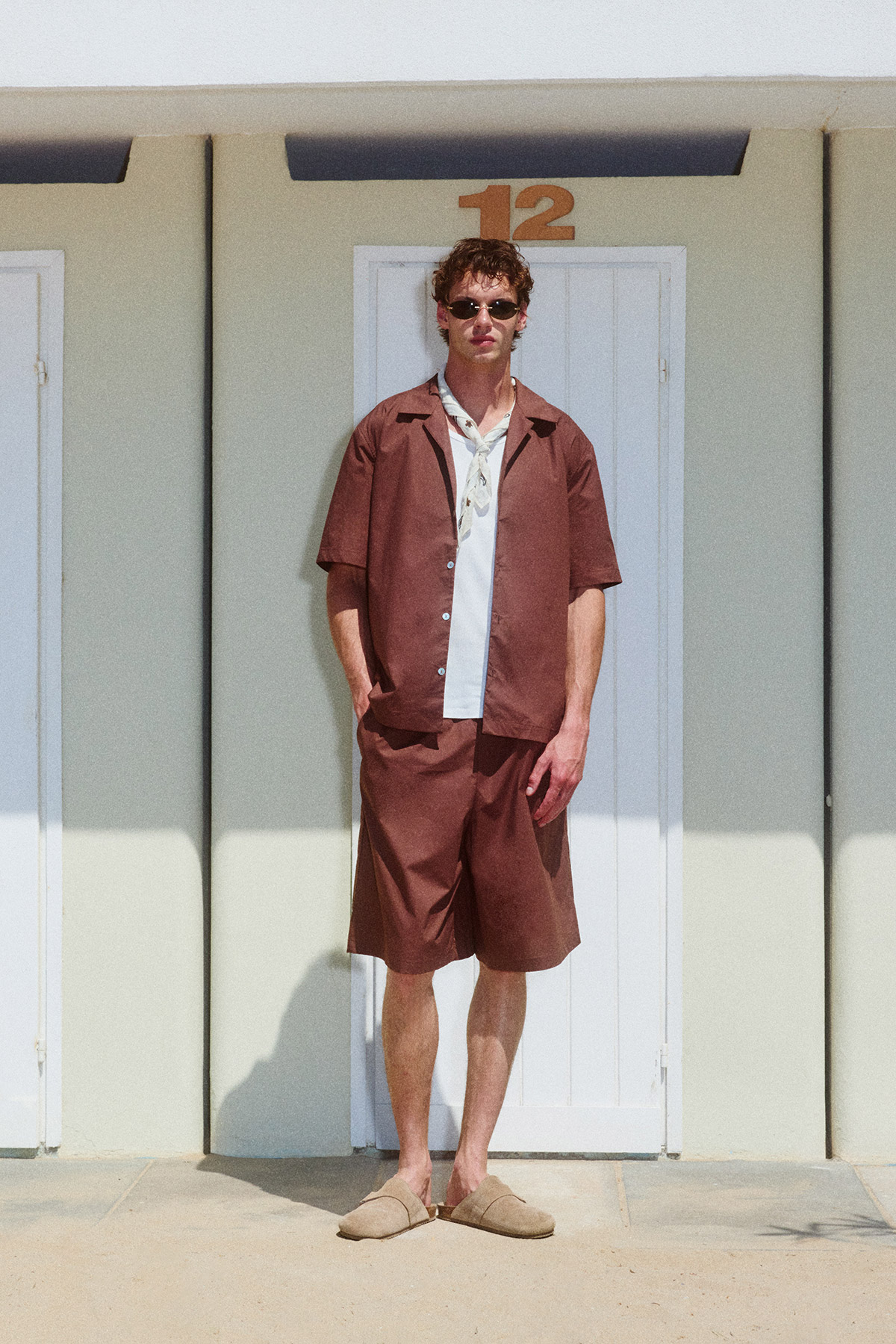 Young man in brown short-sleeve shirt and matching shorts, sunglasses and sandals, standing before a white beach-hut door numbered 12