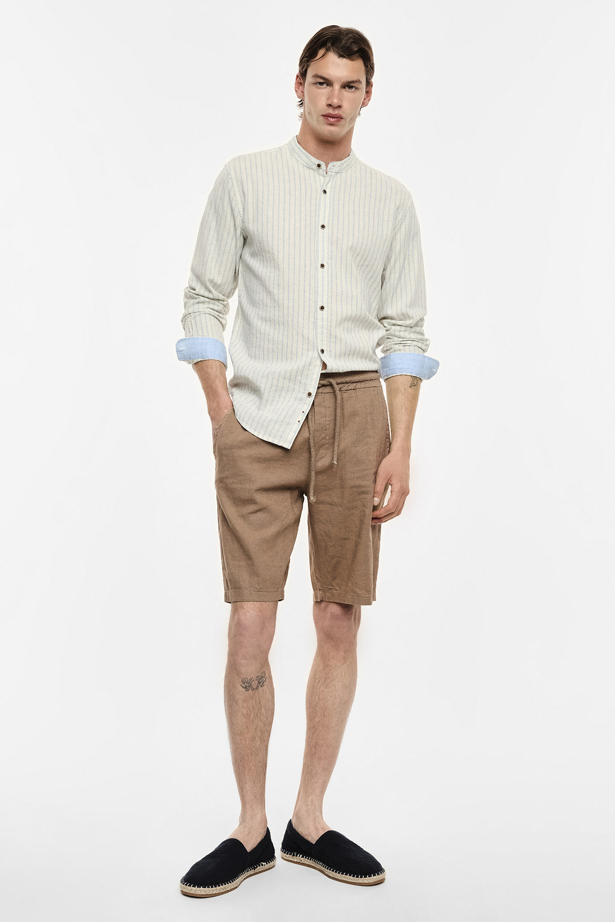 Man wearing cream striped mandarin-collar shirt, brown drawstring shorts and black espadrilles on white background
