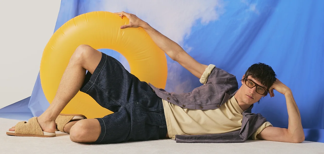 Giovane uomo con occhiali sdraiato sul pavimento che tiene un anello da nuoto giallo, cielo blu con nuvole bianche sullo sfondo, indossa abiti estivi casual.