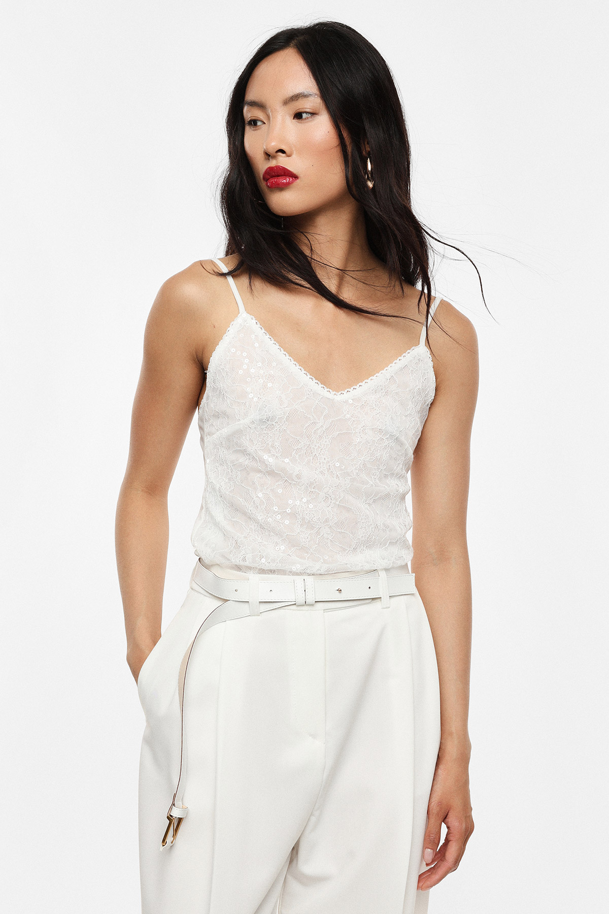 Female model wearing white lace camisole and high-waisted cream trousers, red lipstick, long dark hair, hands in pockets