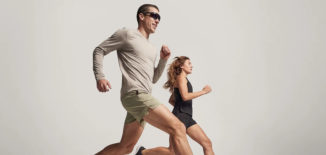 Due adulti, un uomo con pantaloncini e occhiali da sole e una donna con un abbigliamento sportivo nero, che corrono fianco a fianco su uno sfondo semplice.