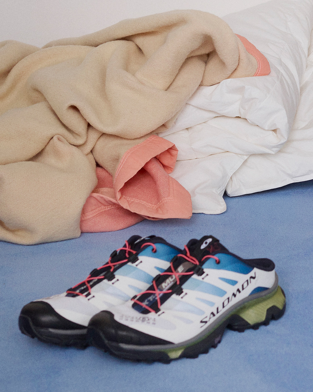 Pair of white-blue Salomon trail running shoes with black soles on a blue floor beside rumpled cream and pink blanket and white pillows