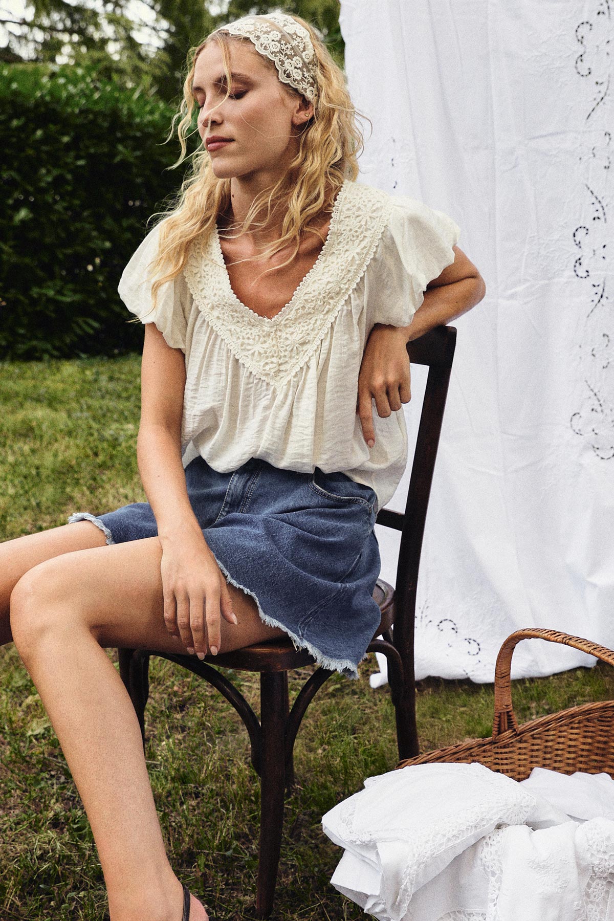 Young woman seated outdoors on a chair wearing a lace headband, embroidered blouse and denim skirt beside a picnic basket