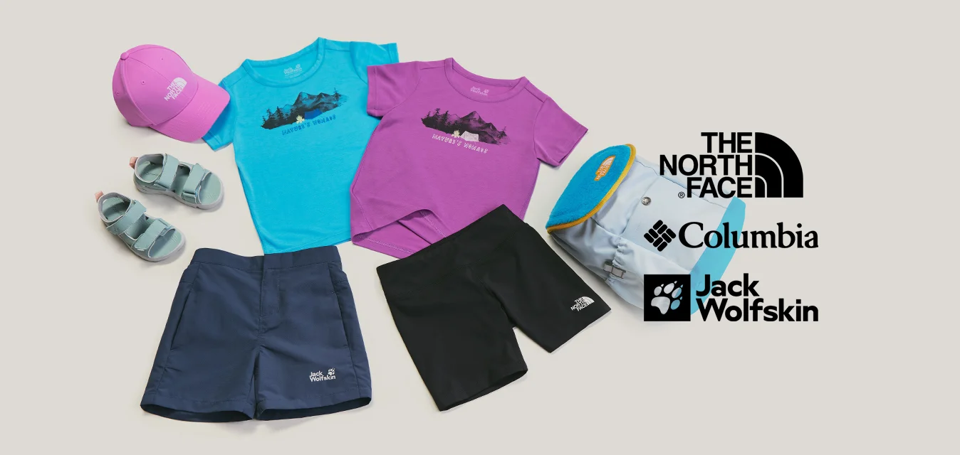 Due t-shirt, due paia di pantaloncini, sandali, un cappellino rosa e uno zaino blu disposti su uno sfondo chiaro.