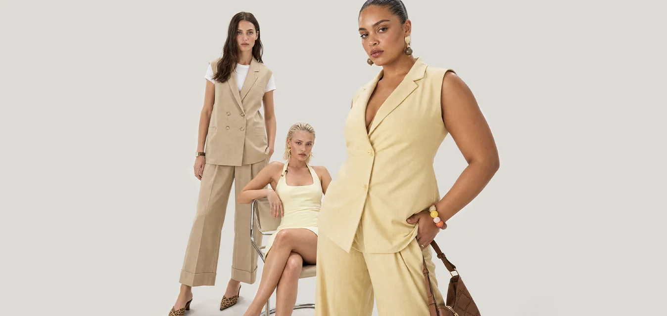 Tre donne in posa con abiti dai toni neutri; una seduta su una sedia indossa una canottiera e una gonna, due in piedi con blazer senza maniche e pantaloni.