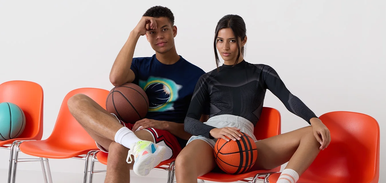 Due persone sono sedute su sedie di plastica arancione, ognuna con una palla da basket in mano. Una indossa una maglietta nera attillata e pantaloni corti, mentre l'altra ha una maglietta colorata e pantaloni corti.