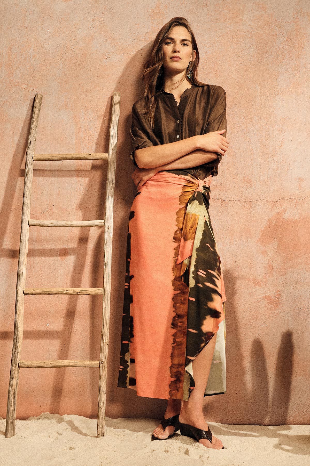 Woman leaning against peach wall beside wooden ladder, wearing brown blouse, coral patterned wrap skirt and sandals