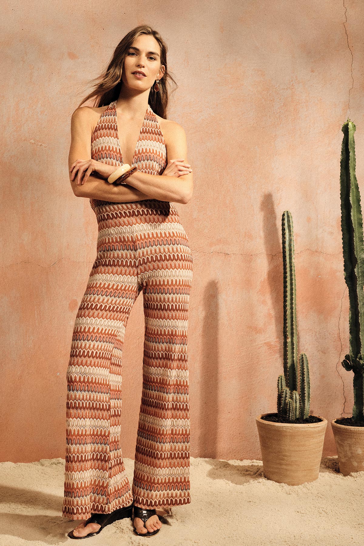 Woman wearing patterned halter jumpsuit and sandals, standing by a terracotta wall with potted cacti