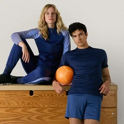 Due persone con abbigliamento sportivo blu posano con una palla arancione su un cavallo da ginnastica in legno contro uno sfondo chiaro e uniforme.