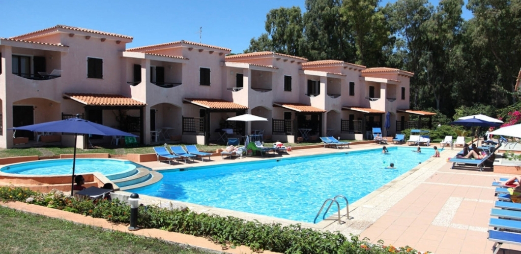 Outdoor swimming pool with sun loungers and umbrellas in front of Mediterranean-style holiday villas surrounded by trees