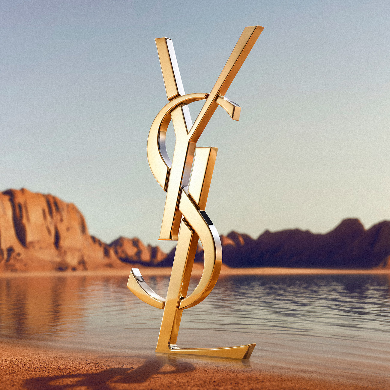 Un monumentale logo dorato YSL che svetta su una riva sabbiosa con montagne e acqua sullo sfondo.