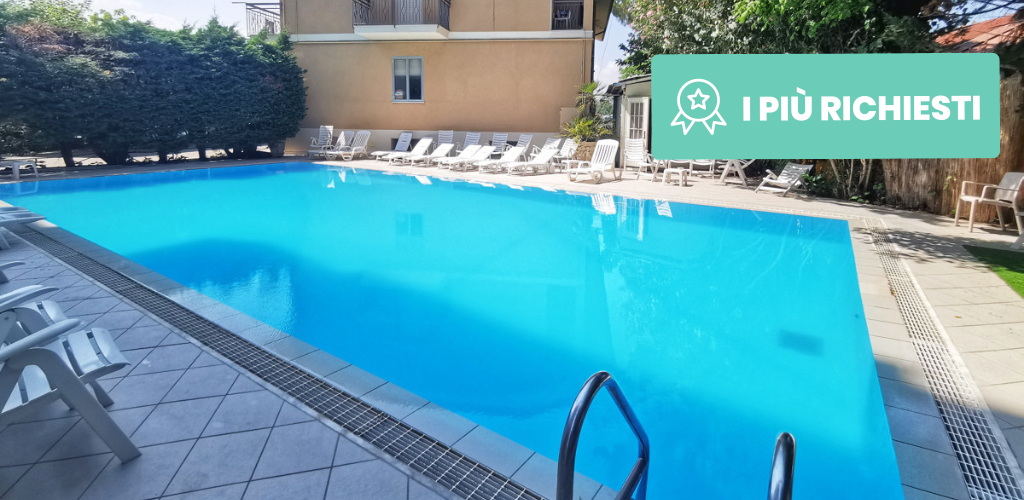 Outdoor hotel swimming pool with clear blue water, metal ladder and sun loungers on a tiled poolside beside a beige building