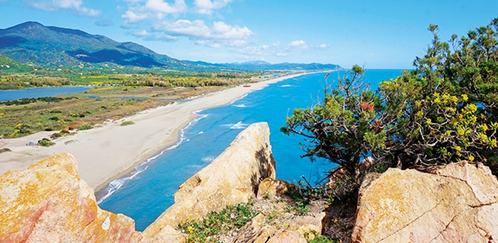 Aerial view of golden cliffs and scrub overlooking a long sandy beach, turquoise sea and distant green mountains under blue sky
