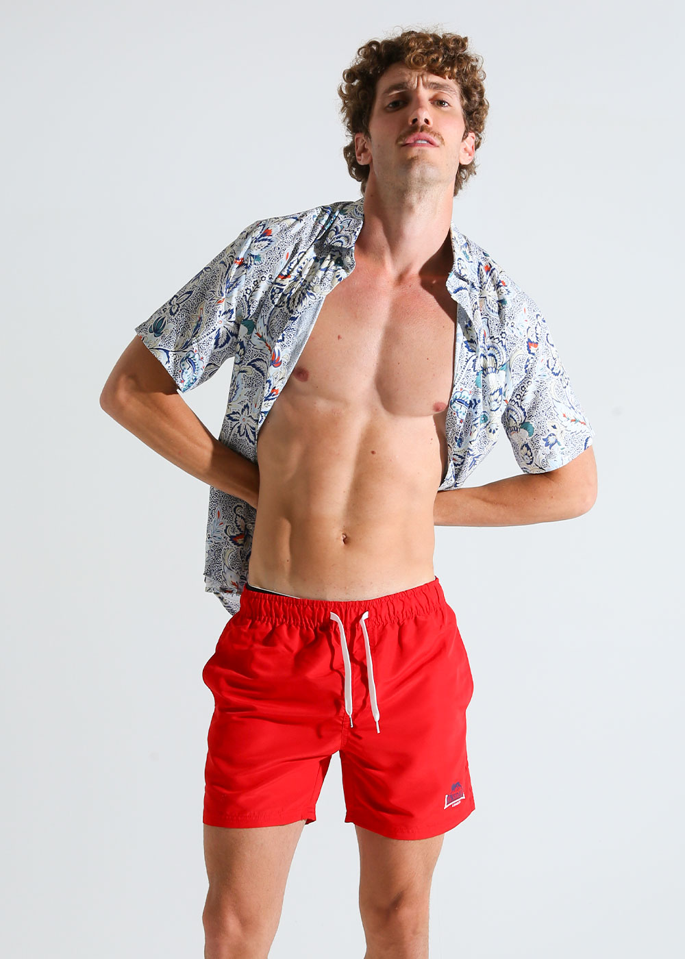 Male model in bright red swim shorts and open patterned short-sleeve shirt, showing toned torso in casual pose