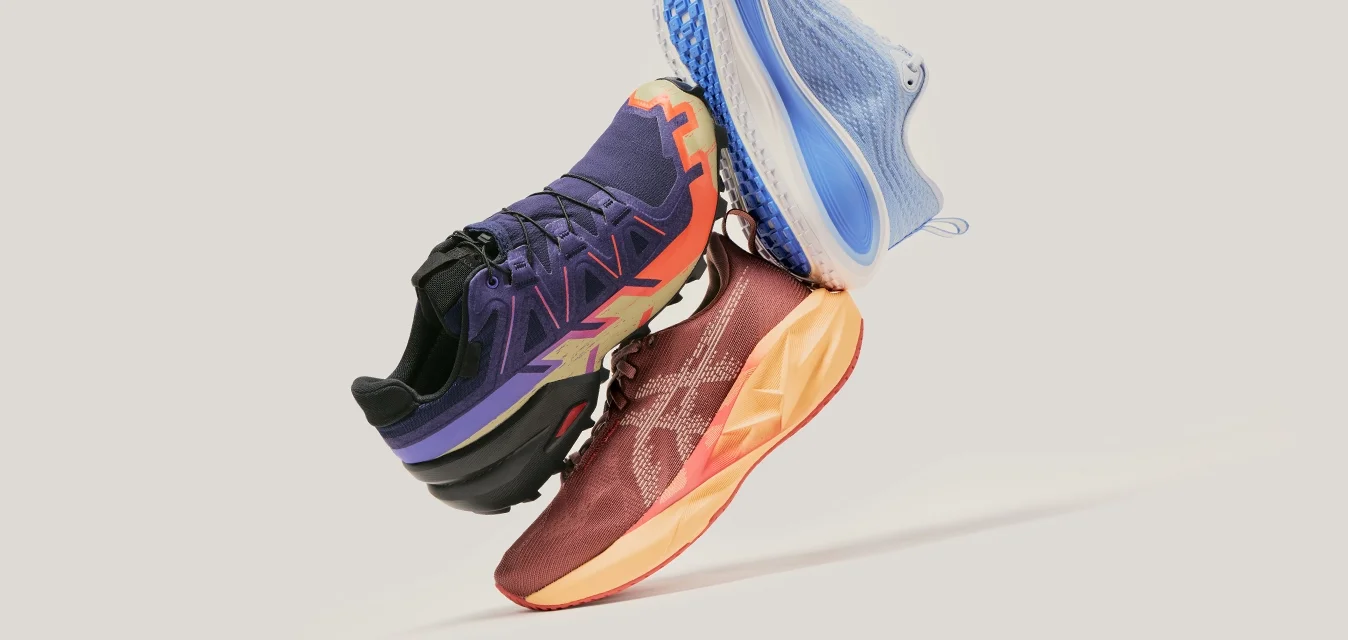 Tre scarpe da ginnastica colorate impilate in aria su uno sfondo grigio chiaro, con design blu, viola e rosso-arancio.