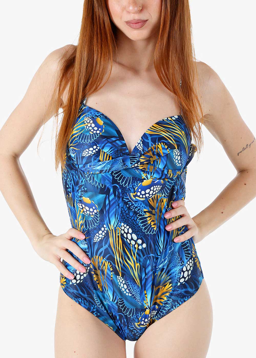 Red-haired woman wearing blue tropical-patterned one-piece swimsuit with seaweed motifs, hands on hips
