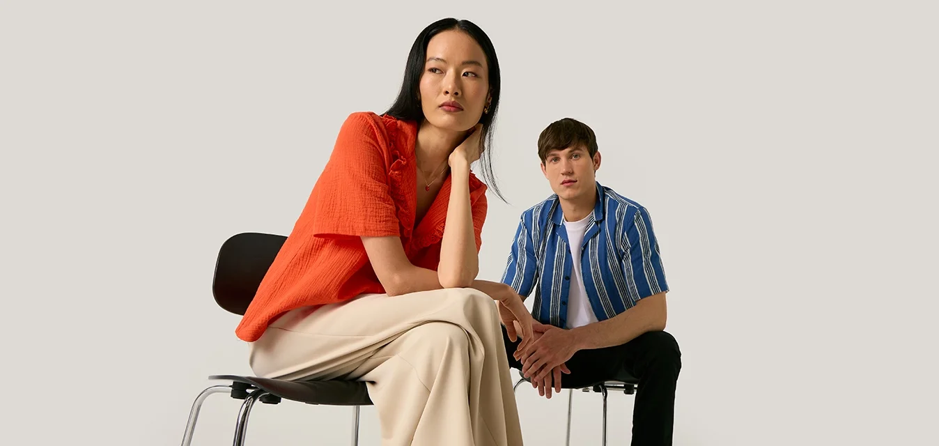 Due persone sedute su sedie davanti a uno sfondo uniforme, una indossa un top arancione e pantaloni beige, l’altra una camicia a righe blu e pantaloni neri.