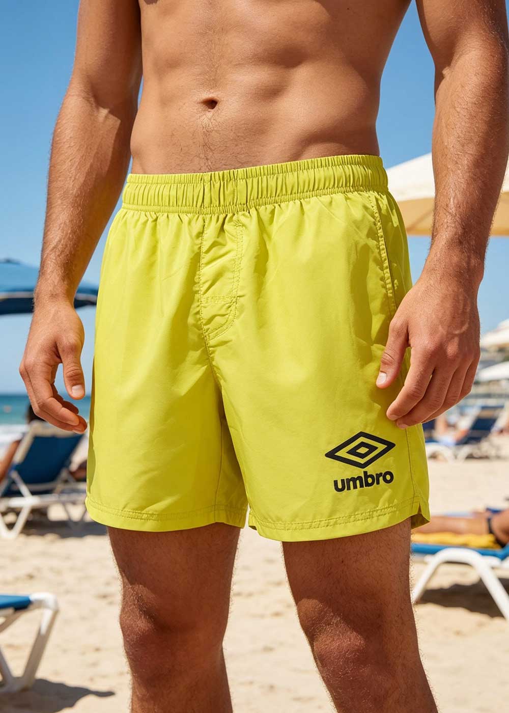 Male torso wearing yellow Umbro swim shorts standing on a sunny beach with sunloungers in background