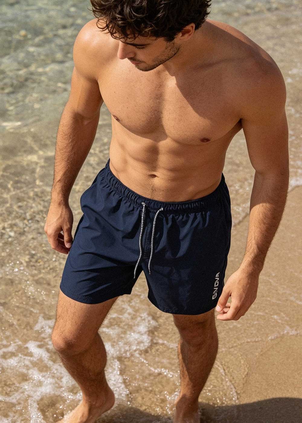 Shirtless man in navy swim shorts standing on a sunlit sandy beach with clear shallow sea water