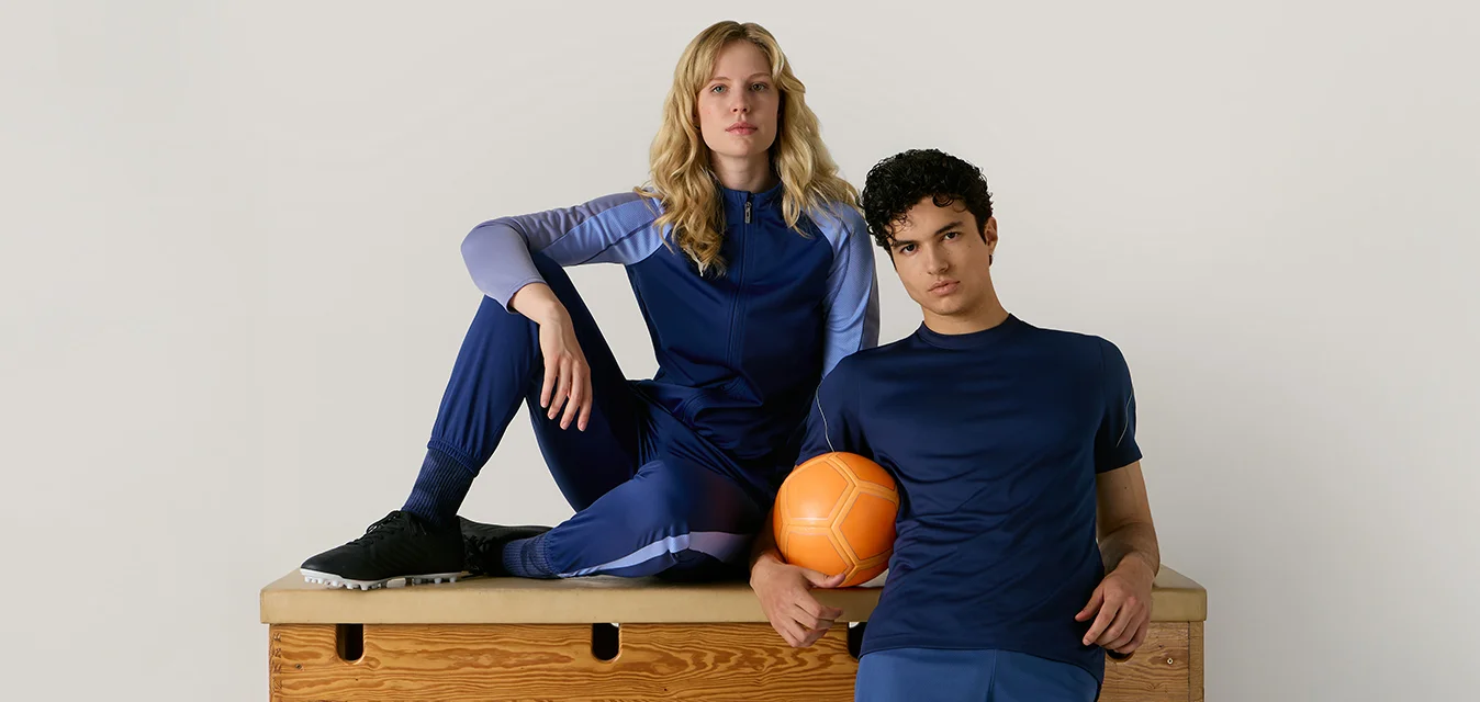 Due persone con abbigliamento sportivo blu posano con una palla arancione su un cavallo da ginnastica in legno contro uno sfondo chiaro e uniforme.