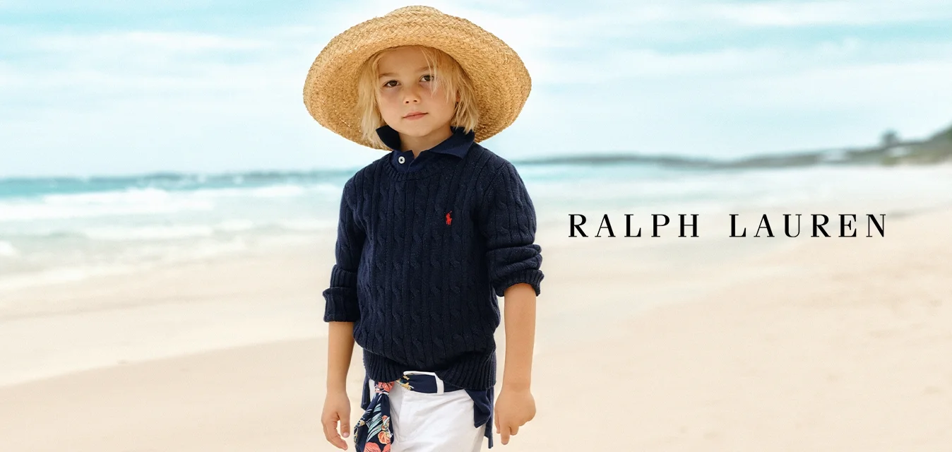 Maglione a collo alto blu navy con lavorazione a trecce, pantaloni bianchi arrotolati e un ampio cappello di paglia. Include una sciarpa a fantasia floreale legata in vita.