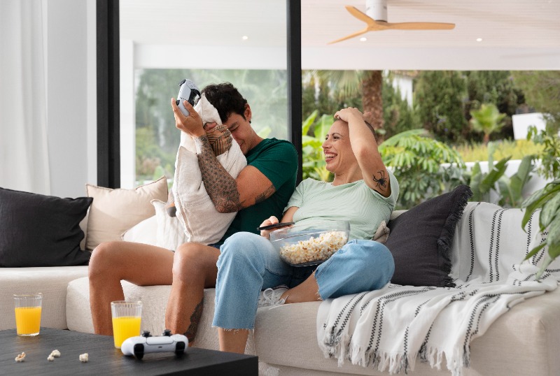 Couple laughing on sofa in living room, playing video games with controllers, bowl of popcorn and glasses of orange juice