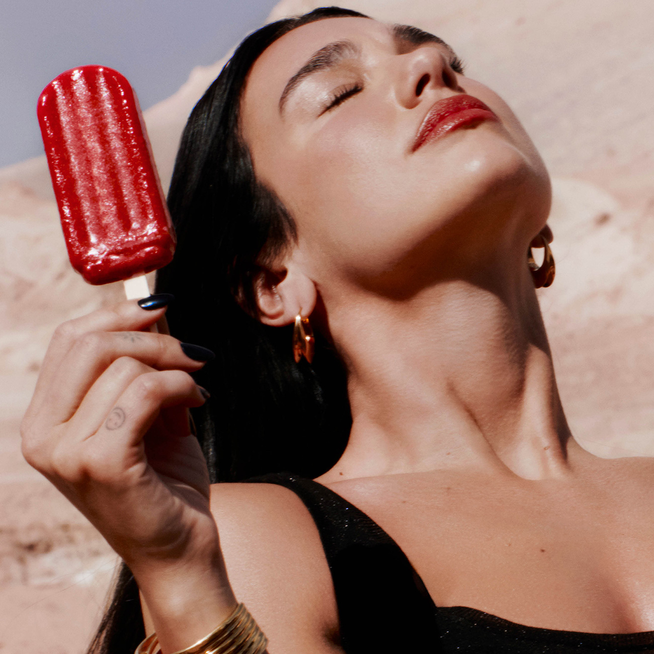 La cantante Dua Lipa inclina la testa all'indietro sotto il sole, indossando orecchini d'oro e tenendo in mano un ghiacciolo rosso.