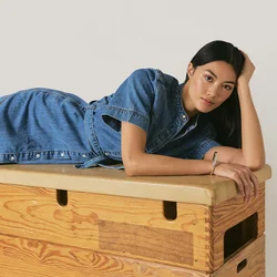Donna con un vestito di jeans blu sdraiata di lato su una cassa di legno da palestra, con la testa appoggiata su una mano, guarda verso la fotocamera.