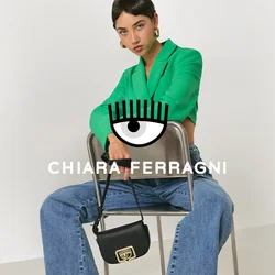 Donna seduta all'indietro su una sedia di metallo, con un blazer verde, jeans larghi blu, tacchi bianchi e sandali con cinturino, mentre tiene una piccola borsa nera.